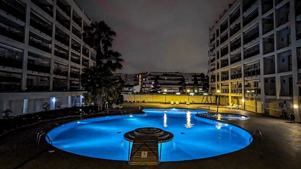 A nighttime swimming pool with a central fountain, surrounded by multi-story buildings.