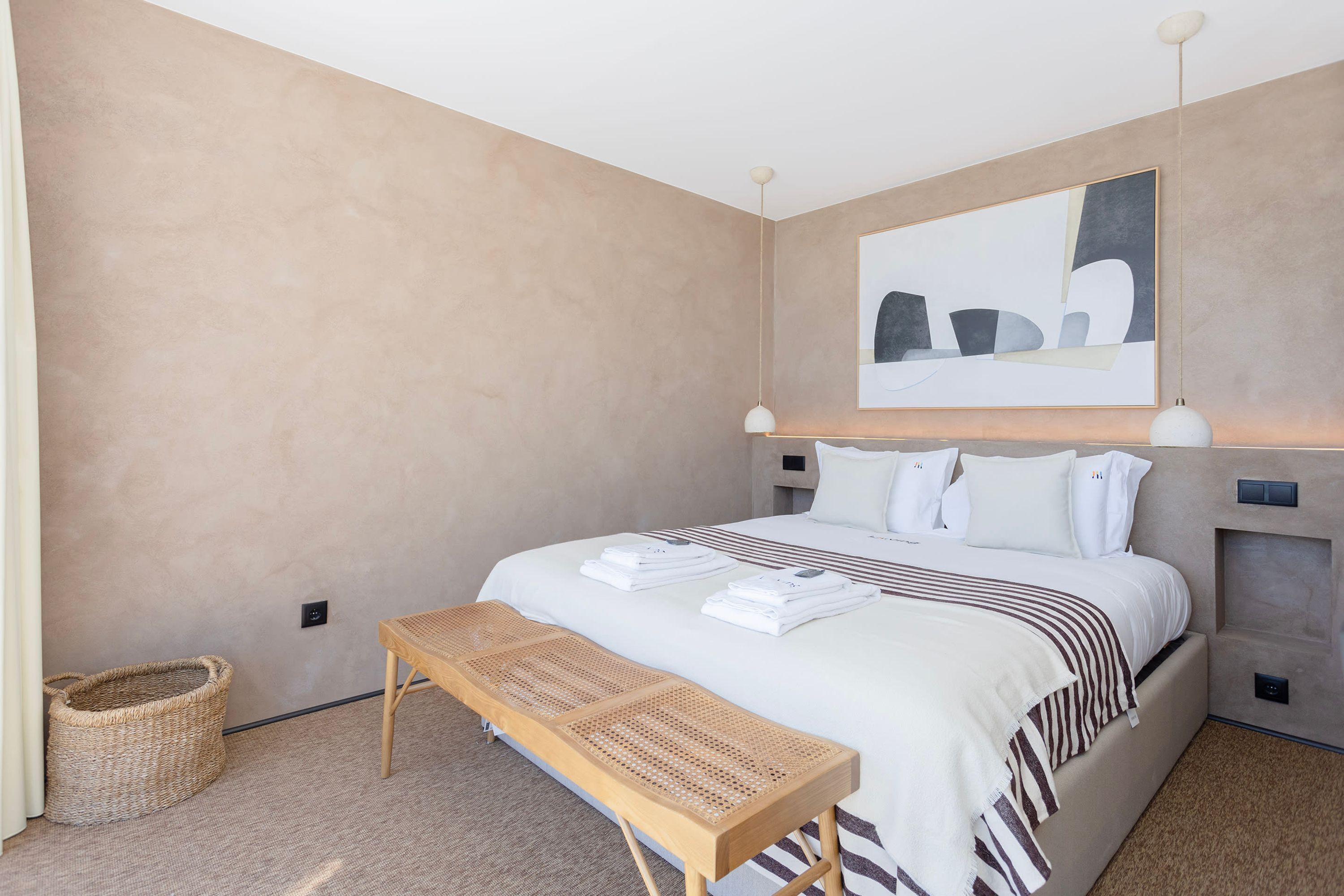 Ein modernes Hotelzimmer mit einem großen Bett, einer Holzbank, einem gewebten Korb und einem großen abstrakten Gemälde an der Wand.
