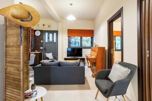 Ein modernes Wohnzimmer mit einer Couch, einem Sessel und einem Fernseher. Es gibt einen Essbereich mit Tisch und Stühlen, und eine durch eine offene Tür sichtbare Küche.