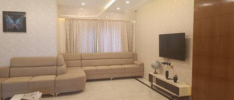 A living room with a beige sectional sofa, a flat-screen TV mounted on the wall, a globe on a small table, and a window with curtains.