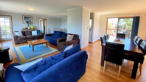 Un salon avec un canapé bleu, un canapé marron, une table basse et une table à manger avec des chaises. Une porte donne accès à un balcon d’où l’on peut admirer des palmiers.