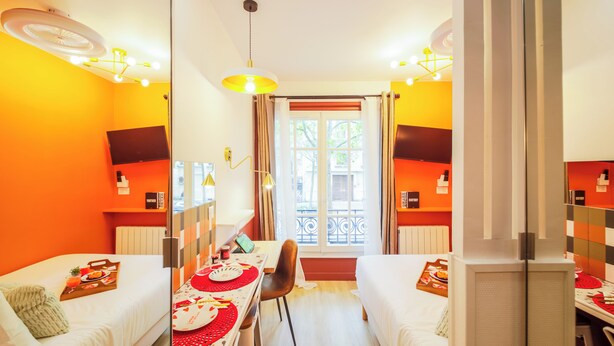 Une chambre d’hôtel avec deux lits simples, une table à manger avec des sets de table rouges, un téléviseur fixé au mur et une fenêtre avec des rideaux.