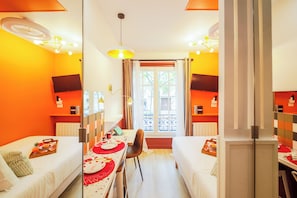 A hotel room with two single beds, a dining table with red placemats, a TV on the wall, and a window with curtains. - WS Montparnasse - Maine (Paris)