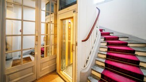Un escalier dont les marches sont recouvertes d’une moquette rouge et qui possède une main courante en bois, menant à un ascenseur.