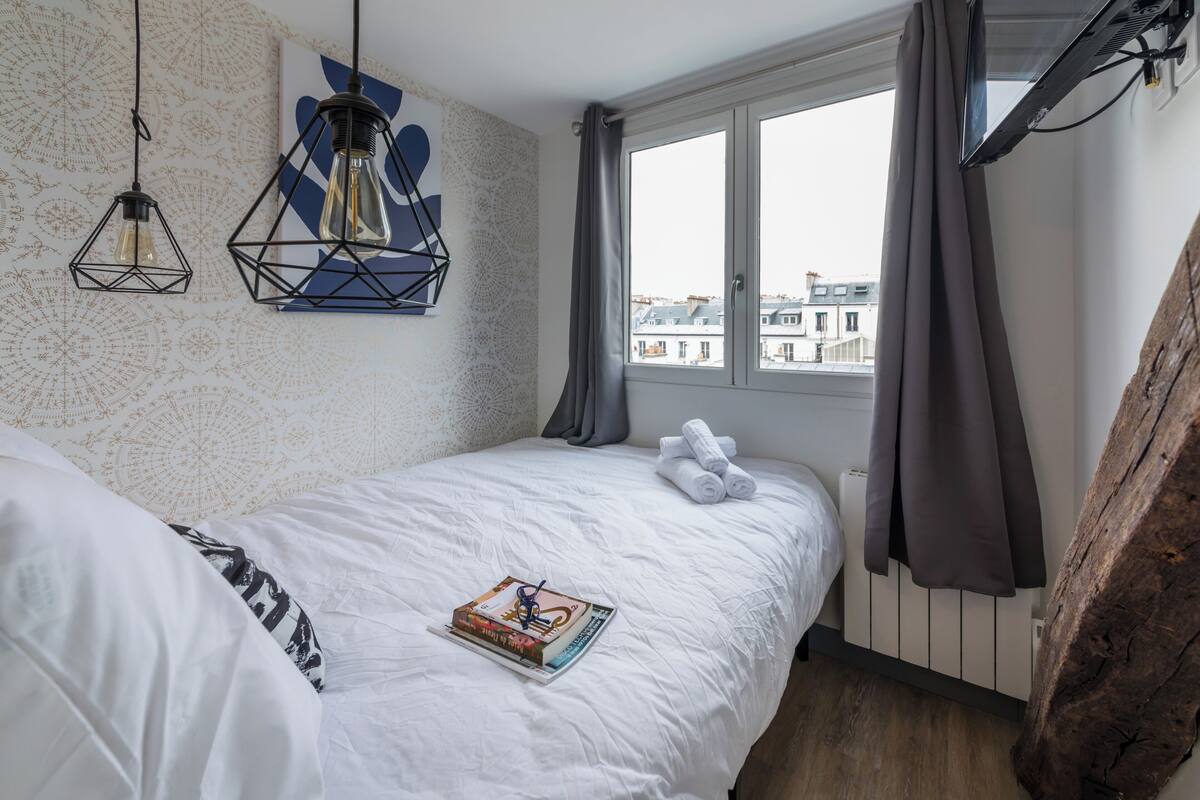 A neatly made bed with a book, a geometric light fixture, and a window with curtains.