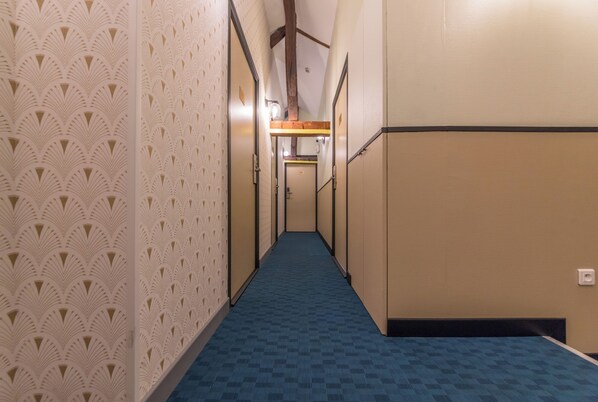 A hotel corridor with patterned walls, a blue carpet, and wooden beams on the ceiling. - WS Saint-Lazare - Lafayette (Paris)