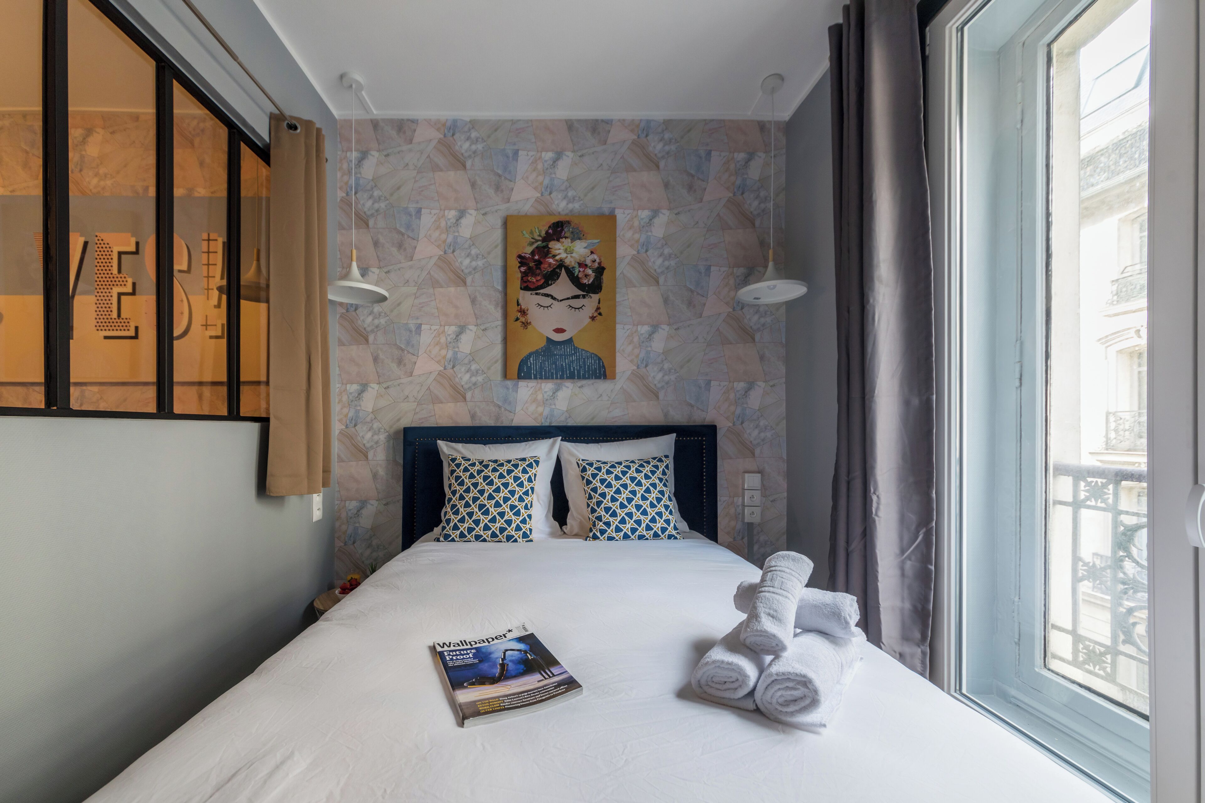 Una cama bien tendida con una revista, toallas enrolladas y un cuadro en la pared.