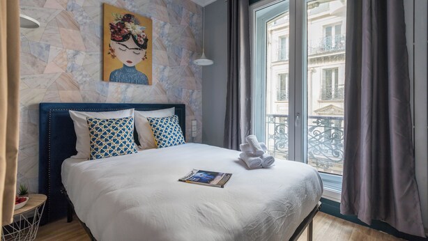 A hotel room with a large bed, a painting on the wall, a window with a view of buildings, and a small table with a plant. - WS Grands Boulevards - Opera (Paris)