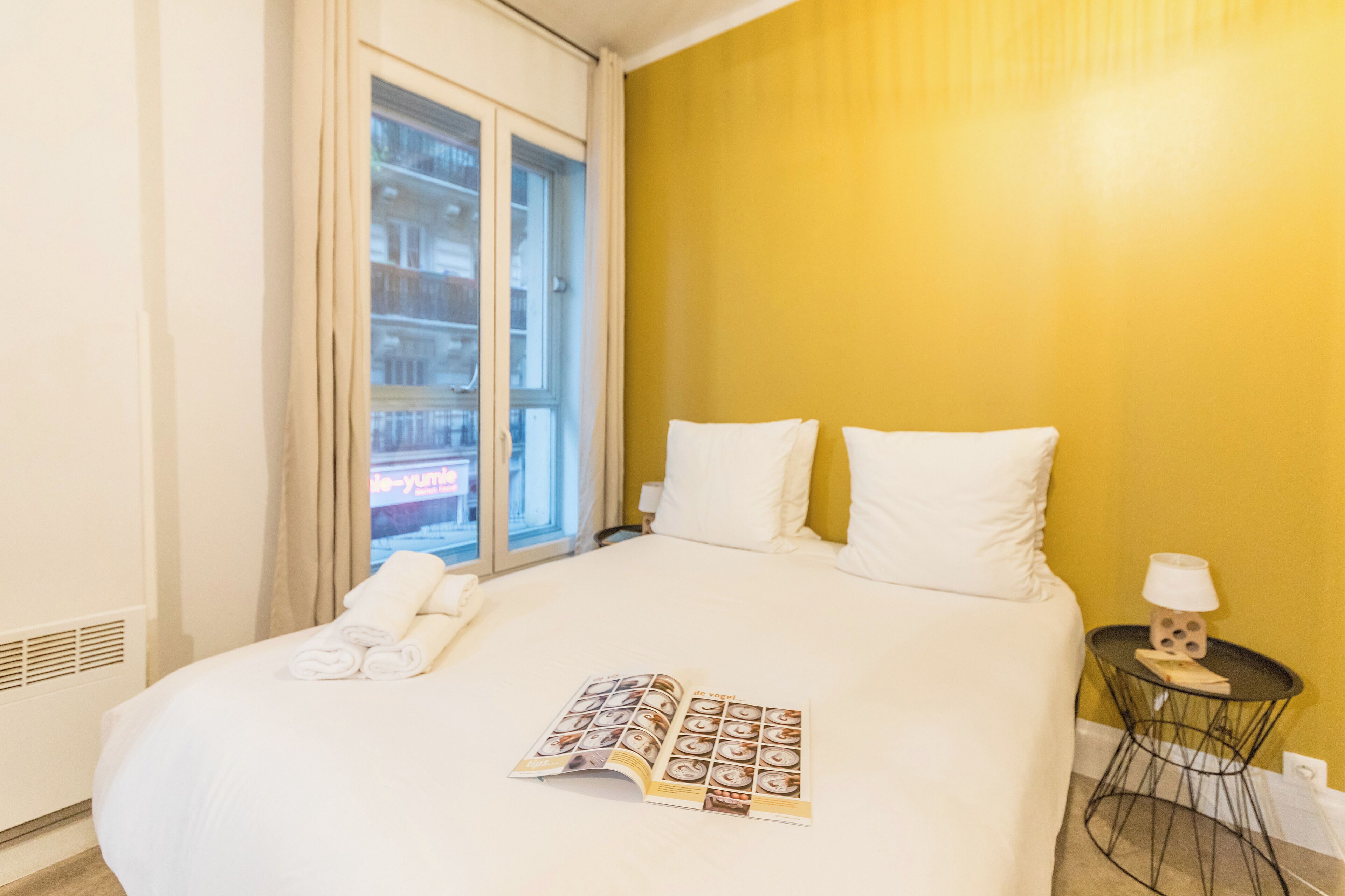 A hotel room with a large bed, a window with curtains, a bedside table with a lamp, and a magazine on the bed.