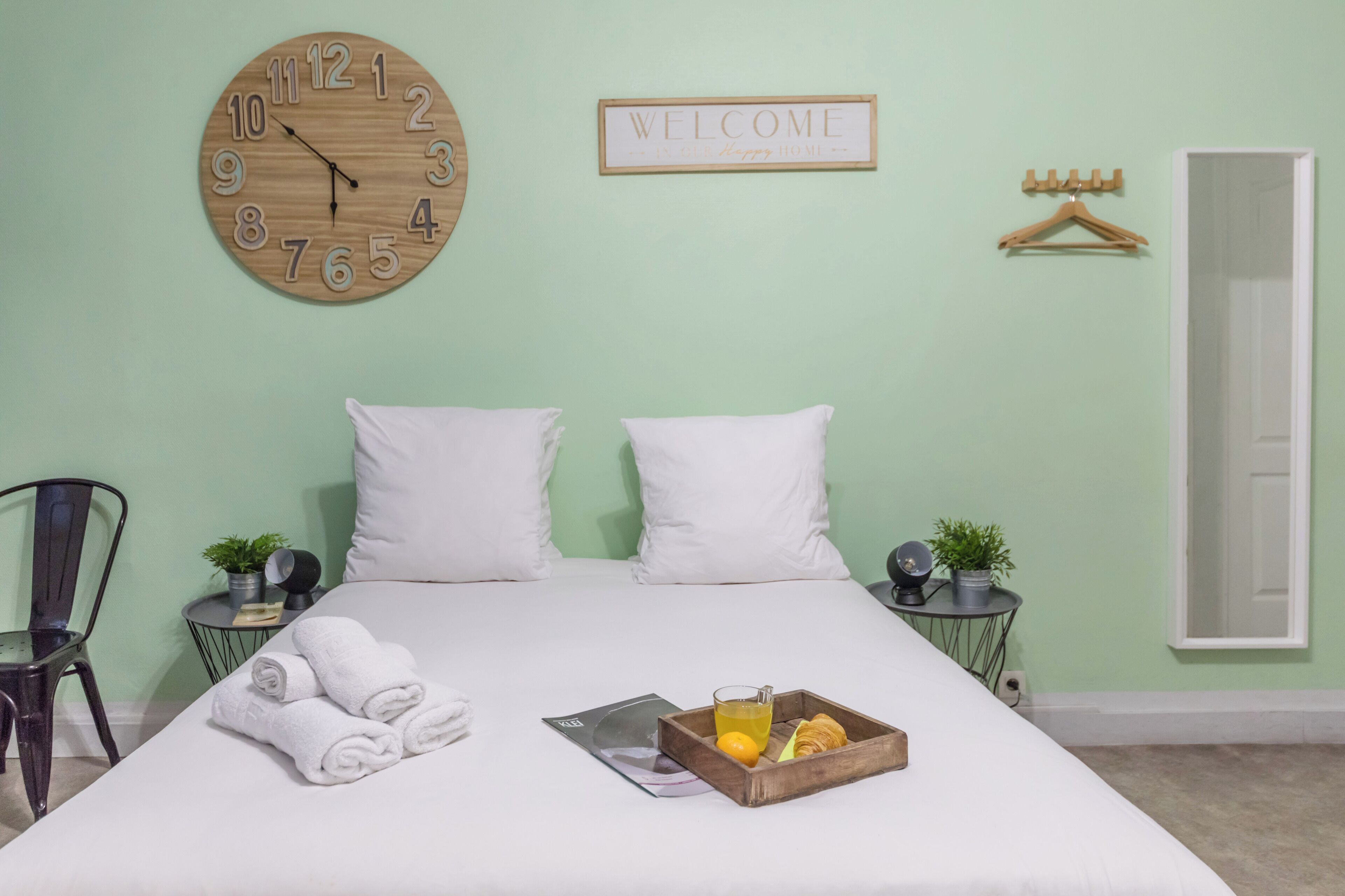 Un letto rifatto con lenzuola bianche, una sveglia in legno, un cartello di benvenuto e un vassoio con prodotti per la colazione.