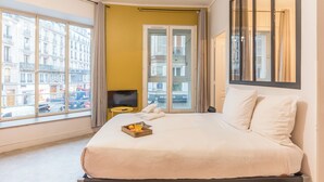 A hotel room with a large bed, a small table with a tray of fruit, a television, and a view of the city through large windows.