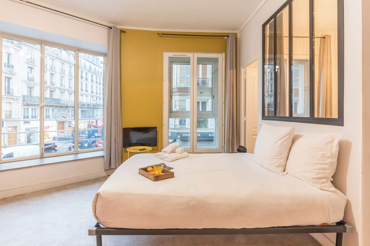 A hotel room with a large bed, a small table with a tray of fruit, a television, and a view of the city through large windows.