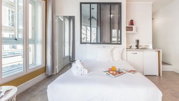 A hotel room with a large bed, a desk with a microwave, and a view of the city through large windows.