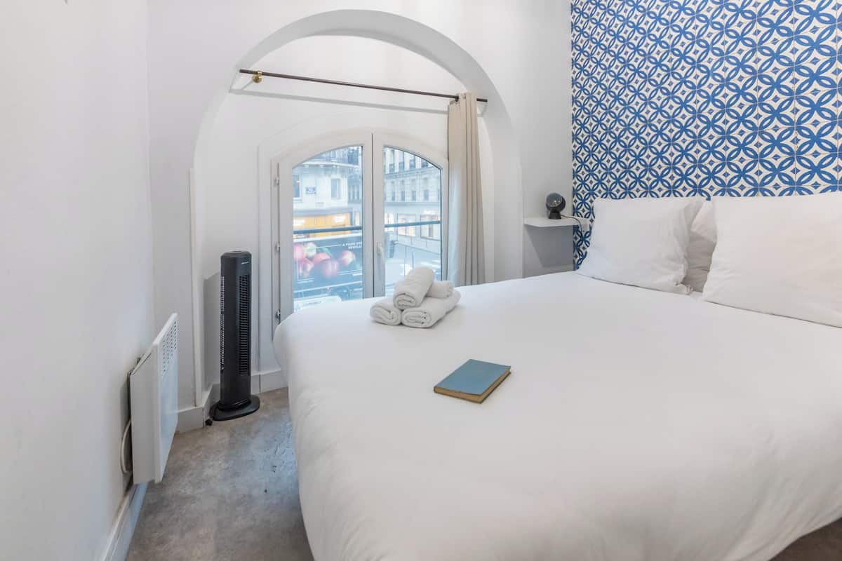 A hotel room with a large bed, a radiator, a standing heater, a window with a view of buildings, and a patterned wall.