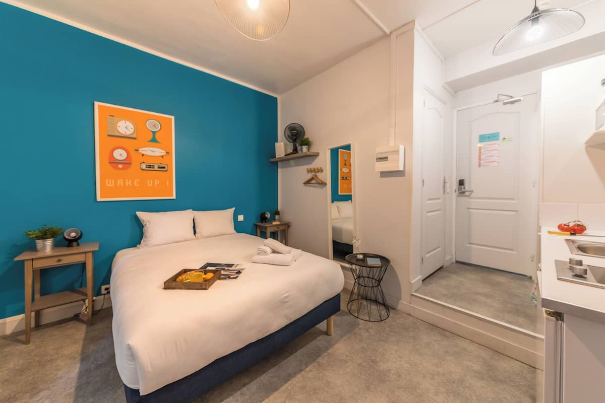 A modern hotel room with a large bed, a bedside table, a small side table, a mirror, a bathroom with a sink and toilet, and a blue accent wall.