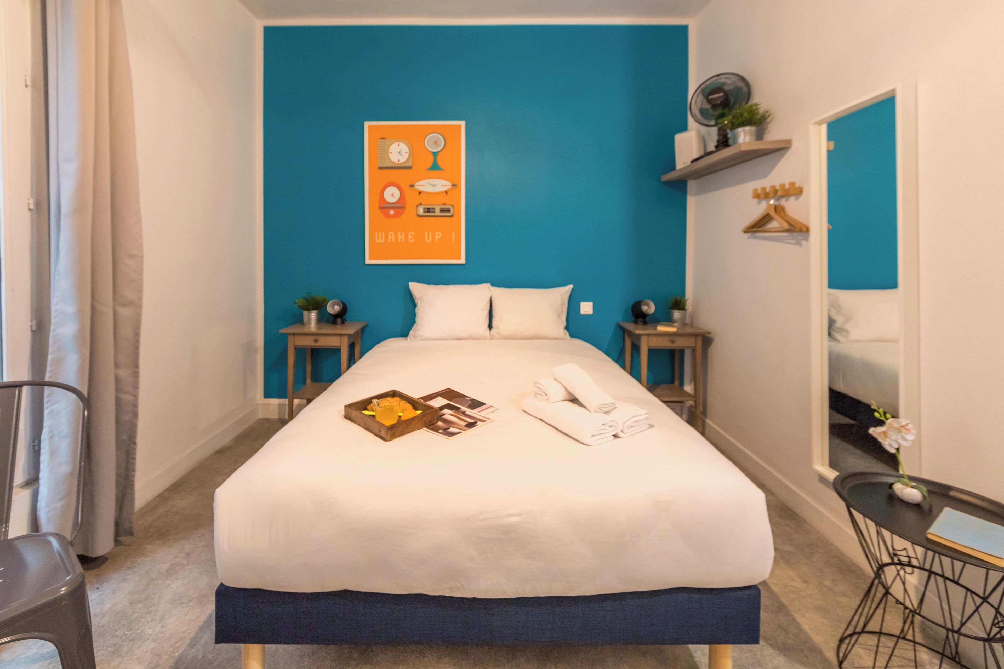 A hotel room with a large bed, a blue accent wall, a bedside table with a clock, a small shelf with a plant, and a mirror on the wall.