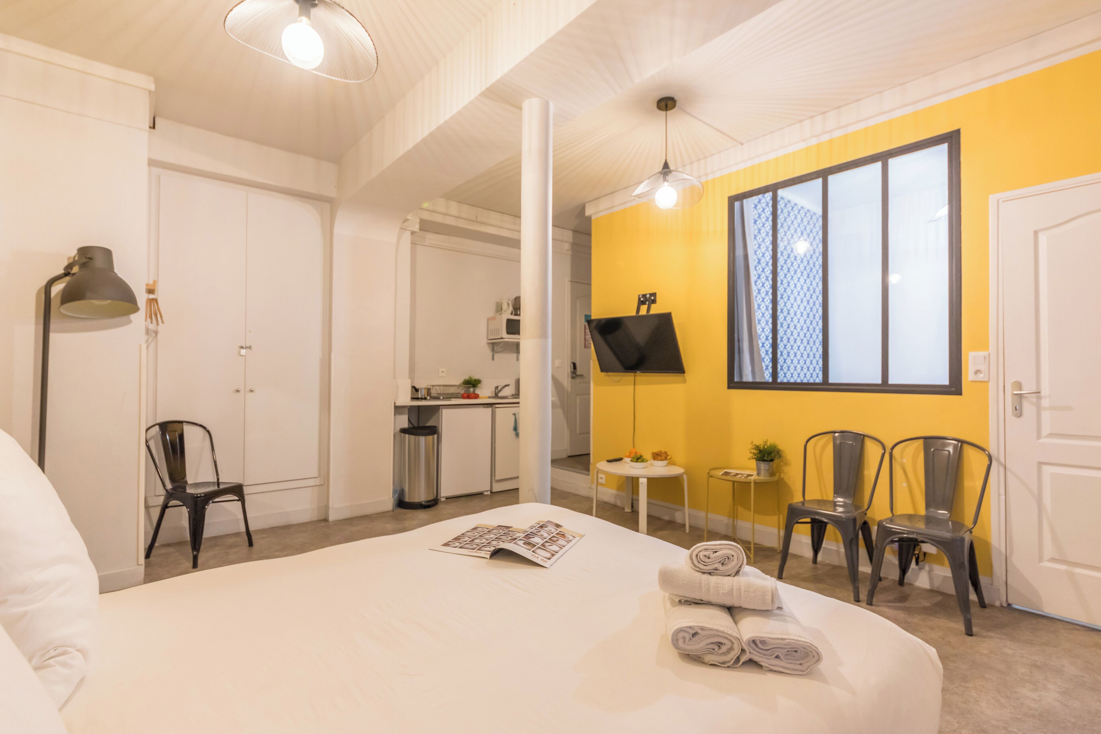 A hotel room with a bed, a desk, a chair, a small table with a plant, and a window with blinds.