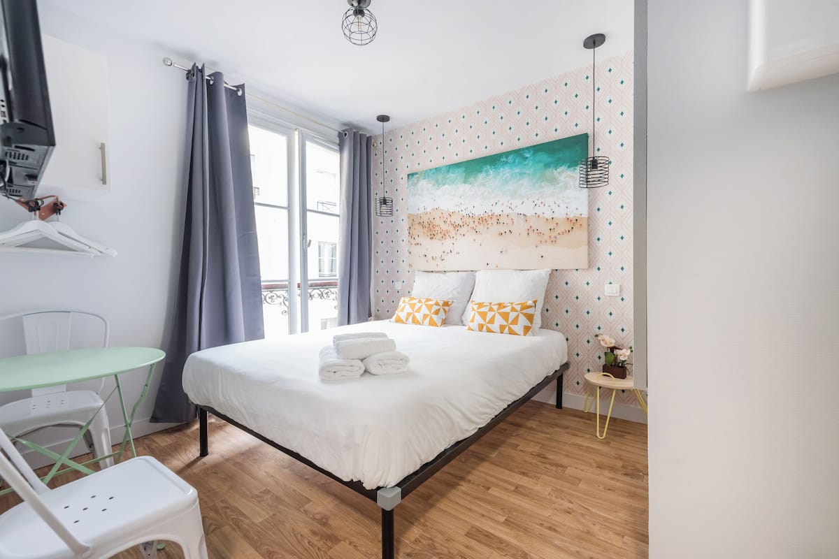 A bedroom with a bed, a table, chairs, a painting, and a window with curtains.
