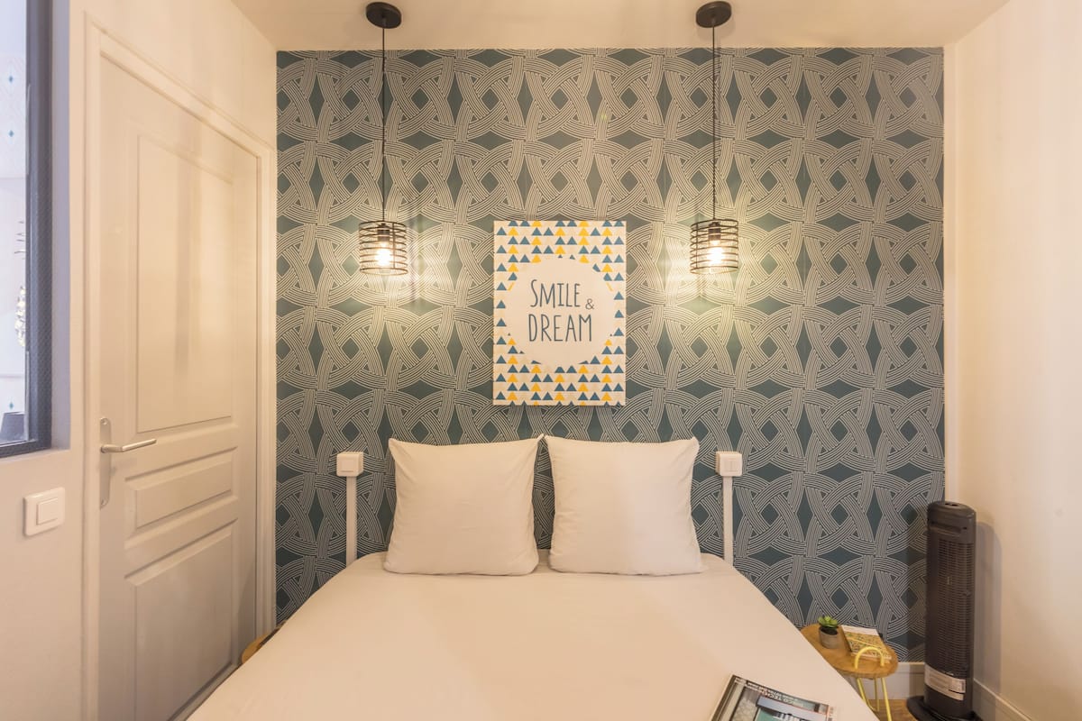 A bedroom with a patterned wallpaper, a bed with two pillows, a framed inspirational poster, two pendant lights, and a small table with a plant.