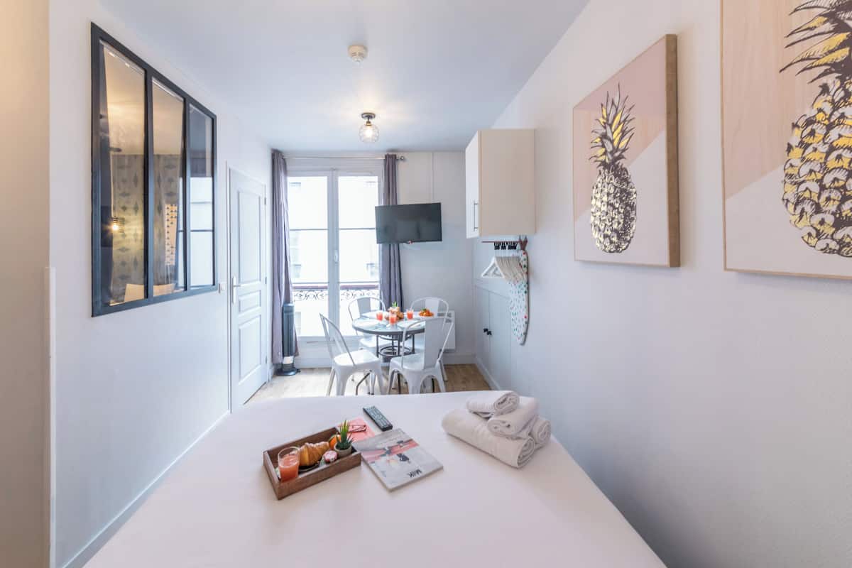 A hotel room with a bed, a dining area with a table and chairs, a TV mounted on the wall, and artwork on the walls.