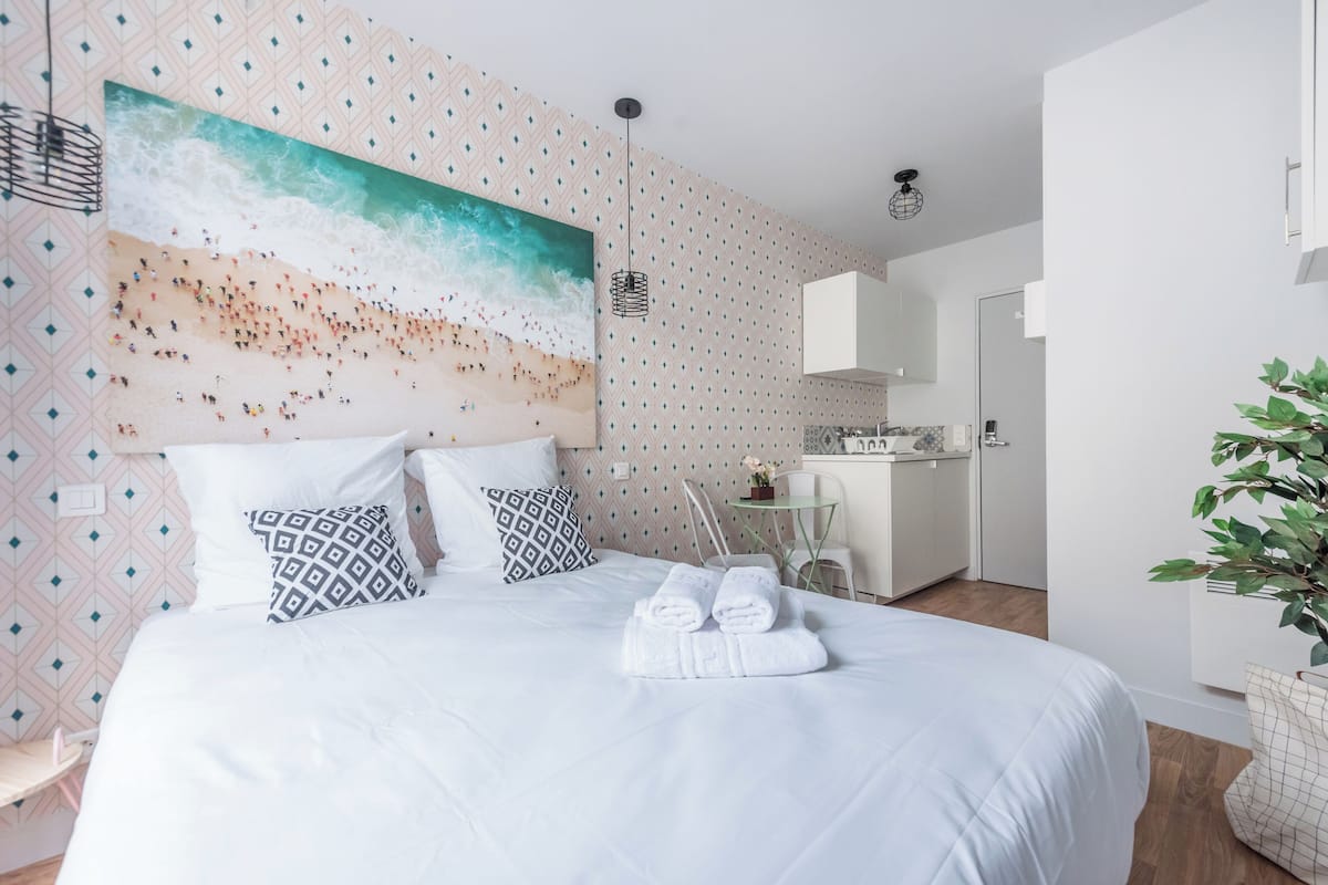A hotel room with a large bed, a painting of a beach scene, a kitchenette, and a dining area.