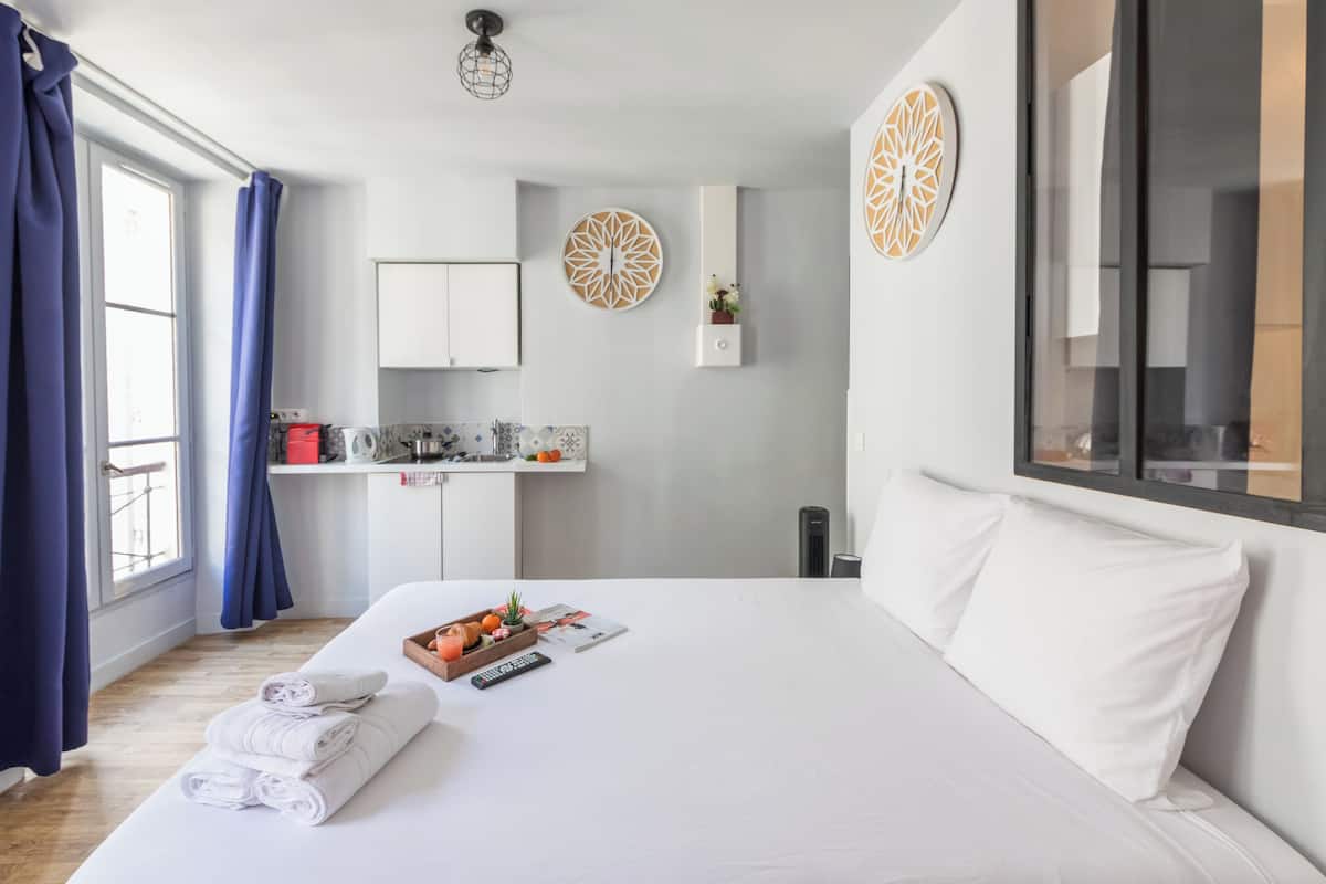 A hotel room with a large bed, a small kitchenette, a window with curtains, and decorative wall art.