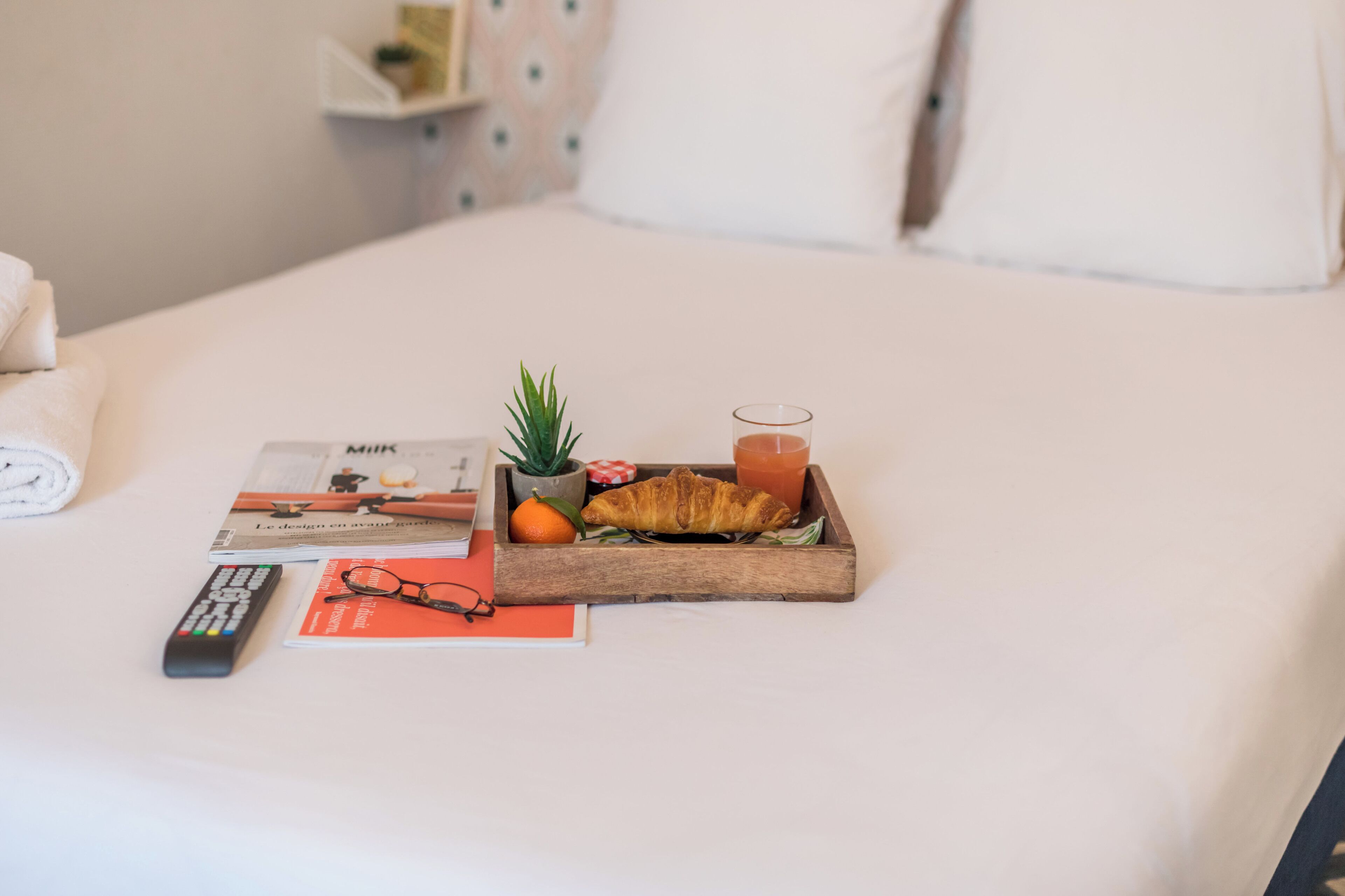 A neatly made bed with a tray containing a croissant, a glass of orange juice, a small plant, and a book.