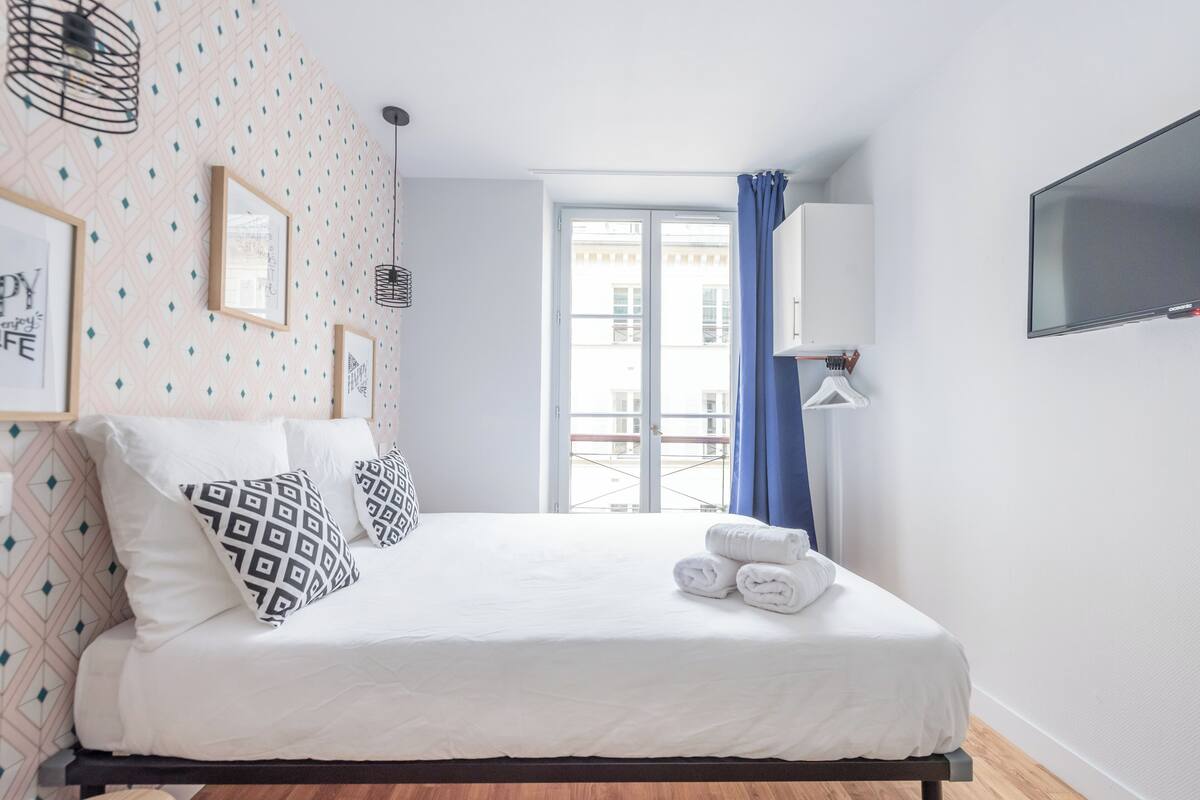 A neatly made bed with patterned pillows, a set of towels, and a window with blue curtains.