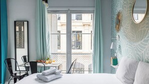 A hotel room with a bed, a dining table with chairs, a balcony with a view of buildings, and a tray with towels and juice. - WS Louvre - Etienne Marcel (Paris)