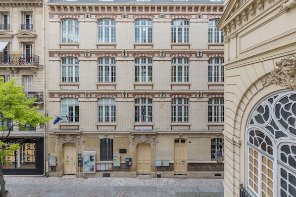 Um edifício de vários andares com fachada simétrica, janelas uniformemente espaçadas, molduras decorativas e uma entrada central com porta e uma pequena varanda.