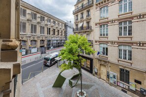 Vista da rua com uma árvore ao centro, edifícios de ambos os lados e um ônibus ao fundo.