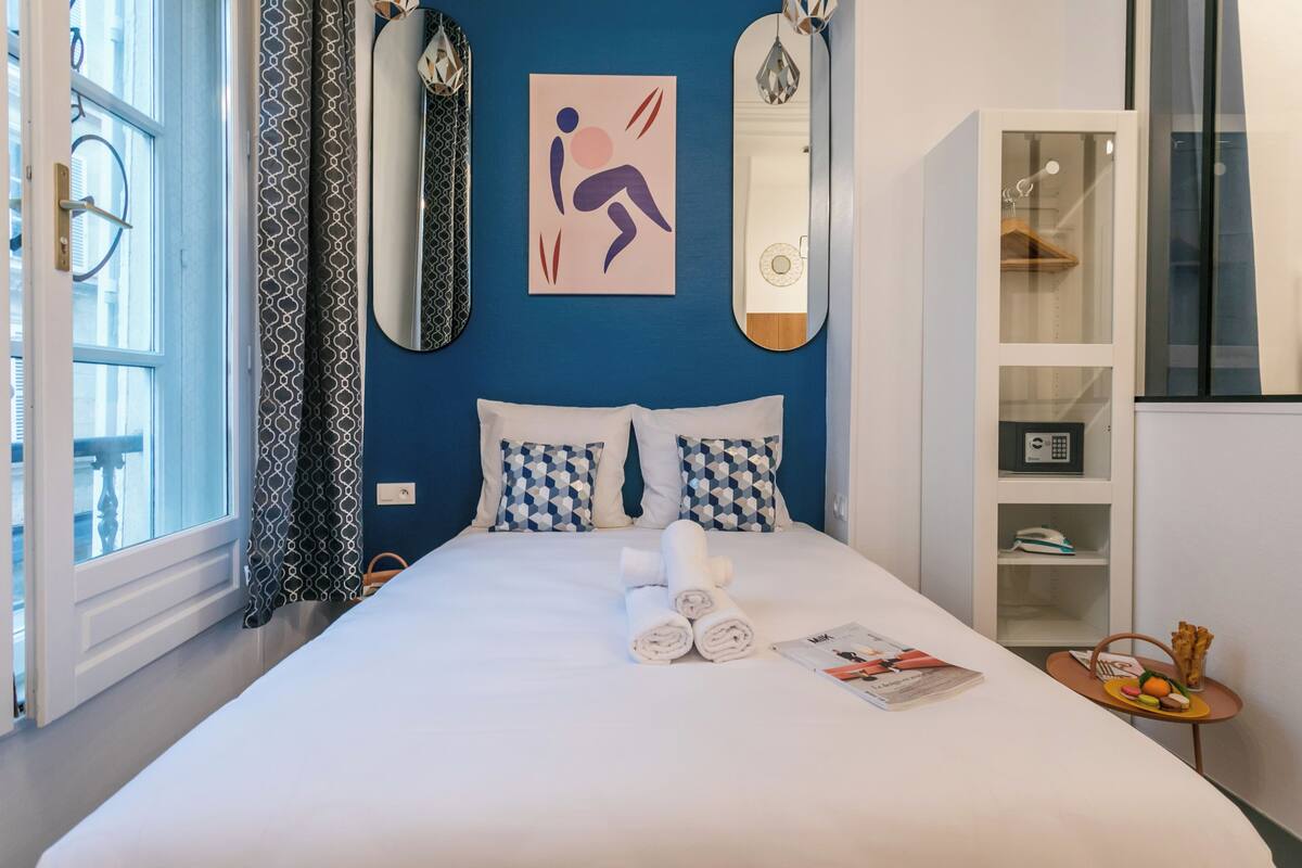 A hotel room with a bed, a window with curtains, a wall-mounted mirror, a towel rack, and a small table with a magazine and snacks.