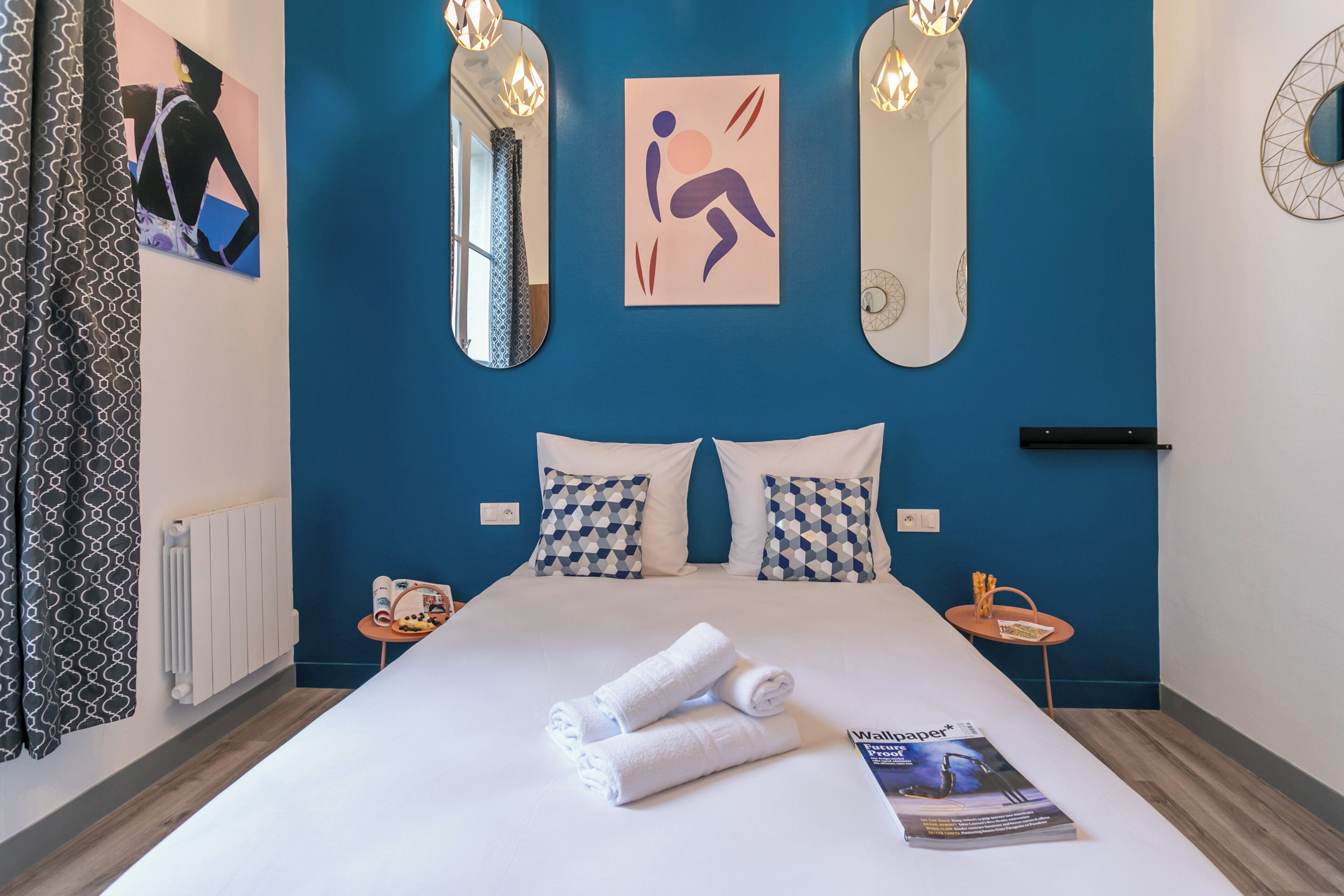 Une chambre d’hôtel avec un grand lit, deux oreillers, une table de chevet avec un magazine, et un mur orné d’un miroir et d’œuvres d’art.