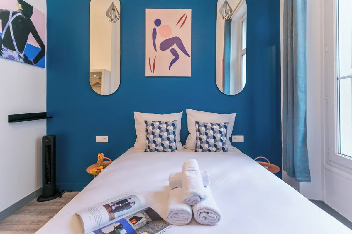 A hotel room with a large bed, two patterned pillows, a wall-mounted mirror, and a window with curtains.