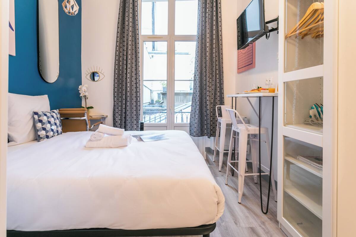 A hotel room with a bed, a desk, a chair, a TV, and a view of the outside.