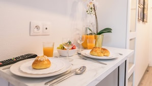 Une table préparée pour le petit-déjeuner avec une télécommande, un verre de jus d’orange, un bol de fruits, un verre d’eau, une assiette avec une pâtisserie et un vase rempli de fleurs.