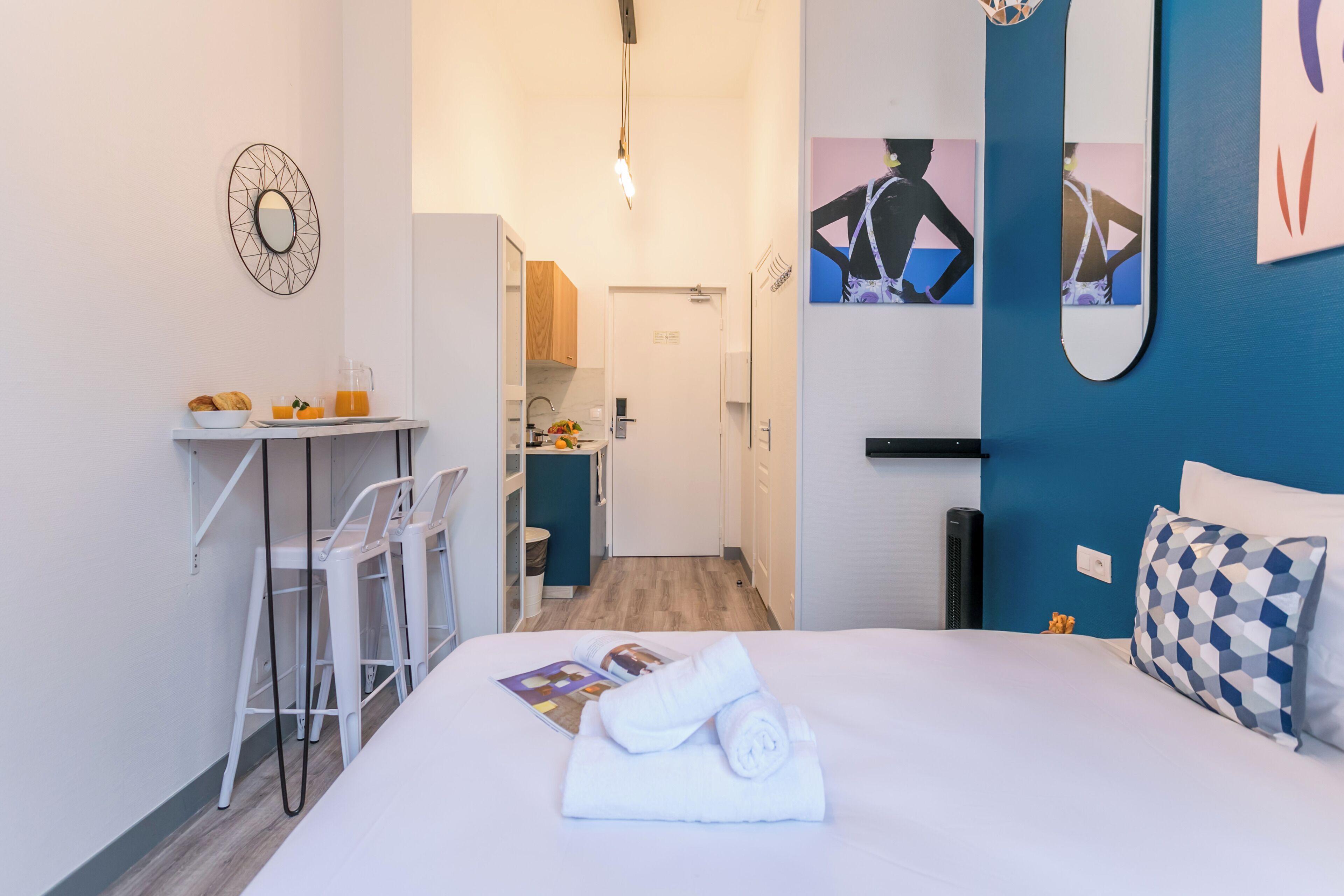 A modern hotel room with a bed, a desk with chairs, a kitchenette, and artwork on the walls.
