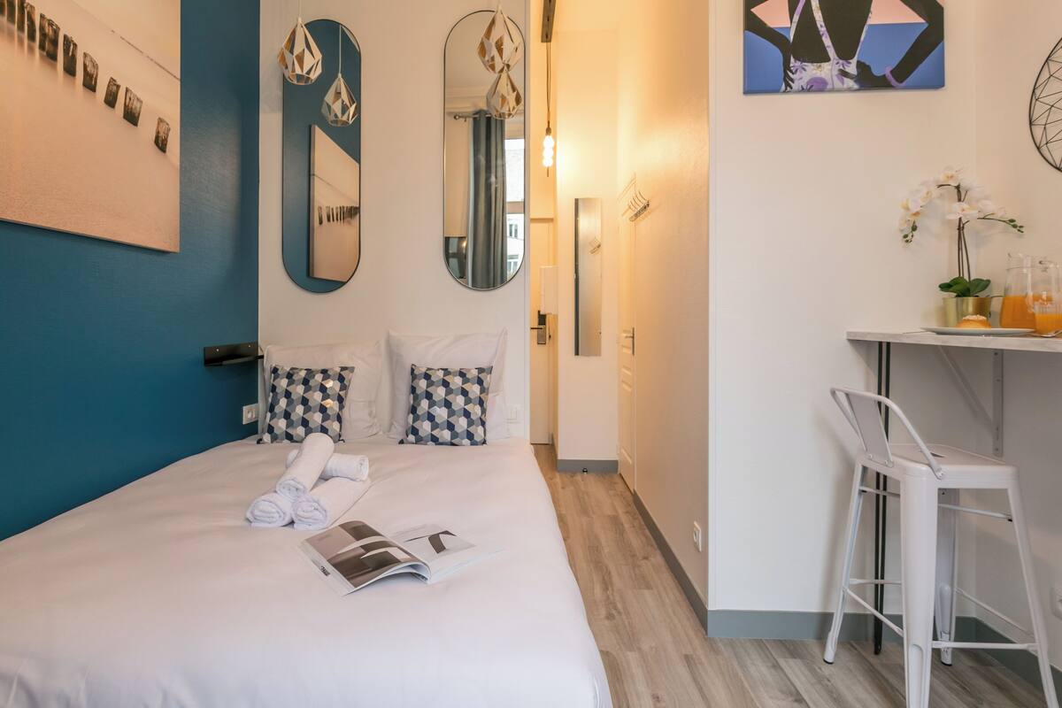 A neatly made bed with pillows, a bedside table with a glass and a plant, and a mirror on the wall.