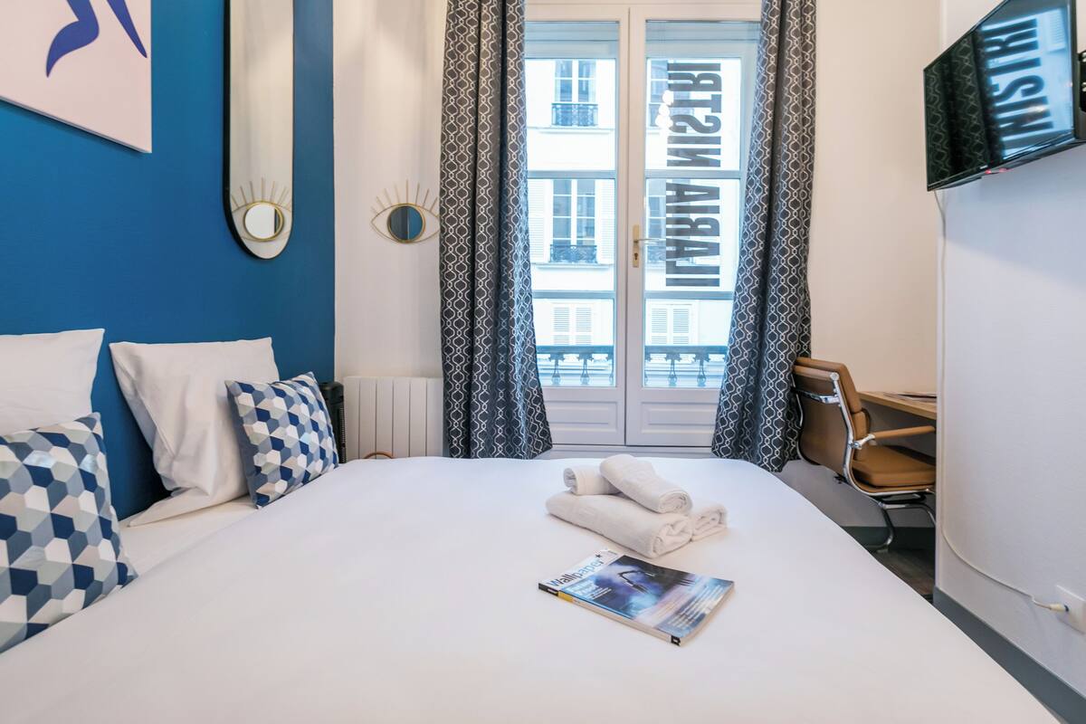 A hotel room with a bed, pillows, a magazine, a chair, and a TV.