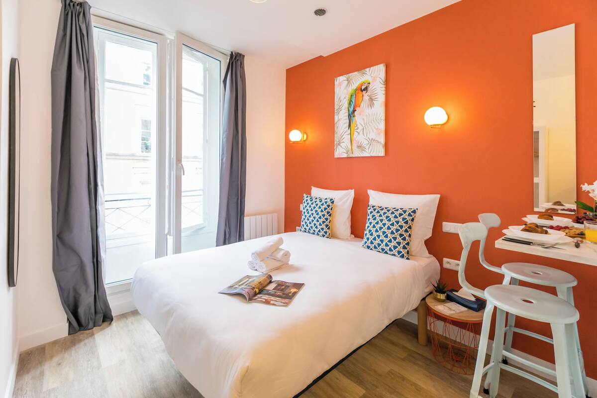 Une chambre d’hôtel avec un grand lit, une petite table avec des articles pour le petit-déjeuner, un miroir et une fenêtre avec des rideaux.