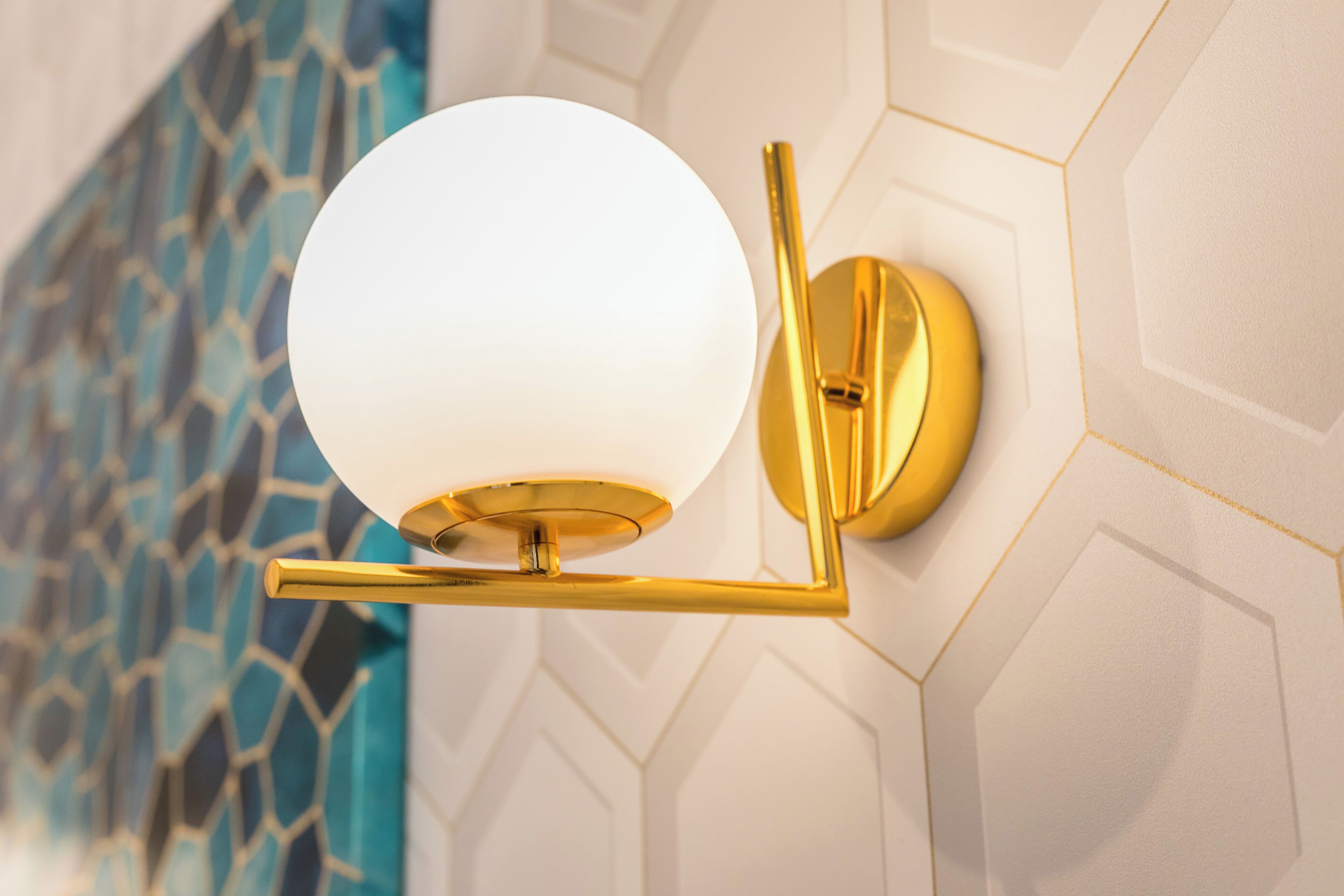 Una lámpara de pared con pantalla de cristal esférica y base dorada, sobre un fondo de pared con diseño geométrico.