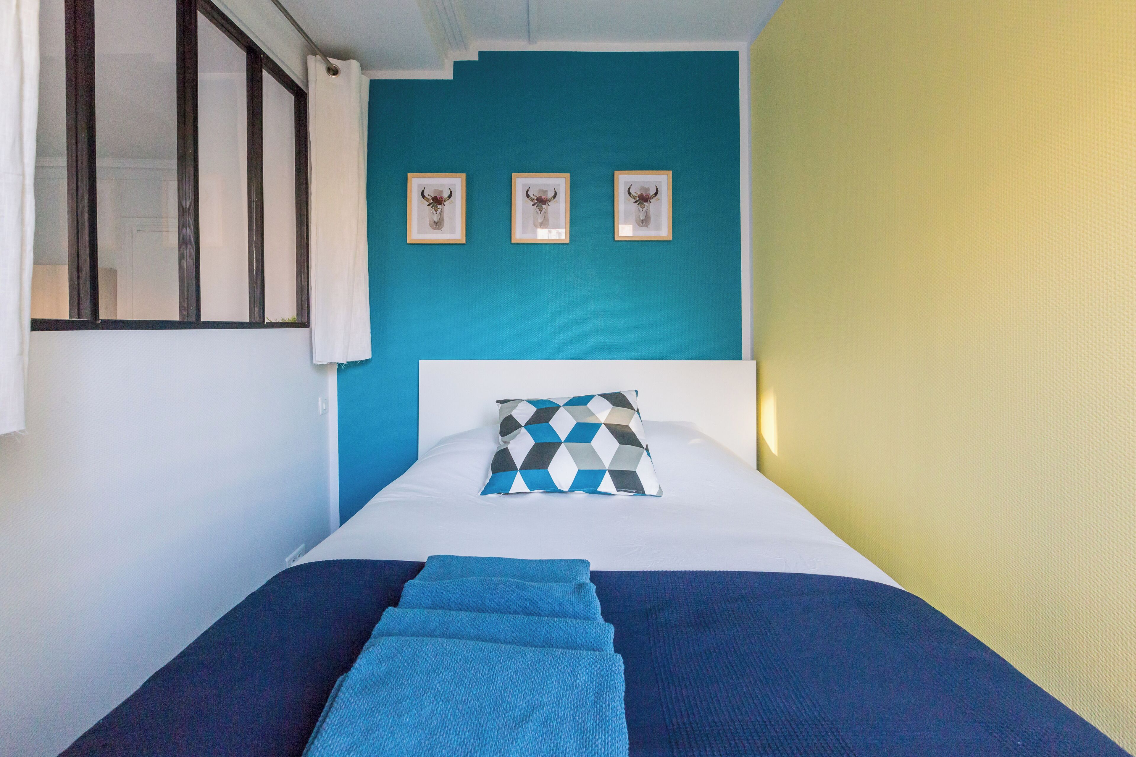 Un lit recouvert d’une couvre-lit bleue et blanche, un coussin à motifs géométriques et trois têtes d’animaux encadrées accrochées au mur.