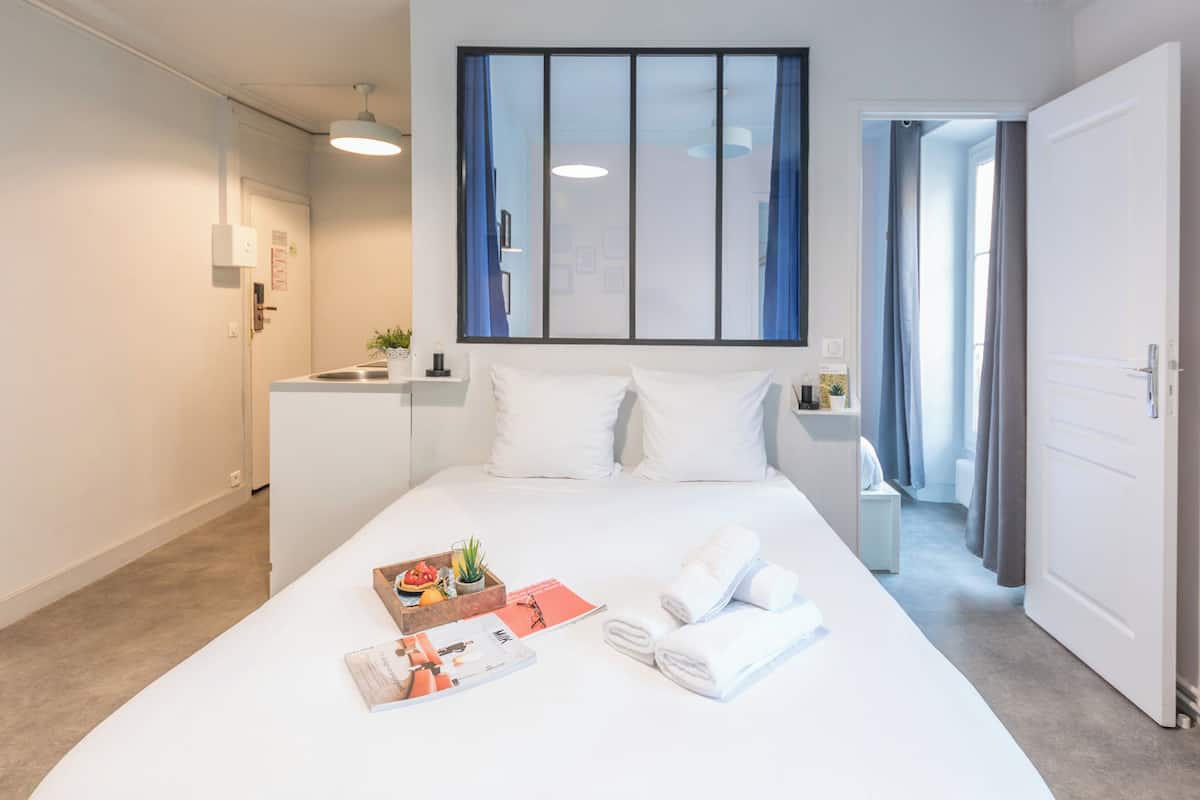 A hotel room with a large bed, a small desk, a window with curtains, and a door leading to another room.