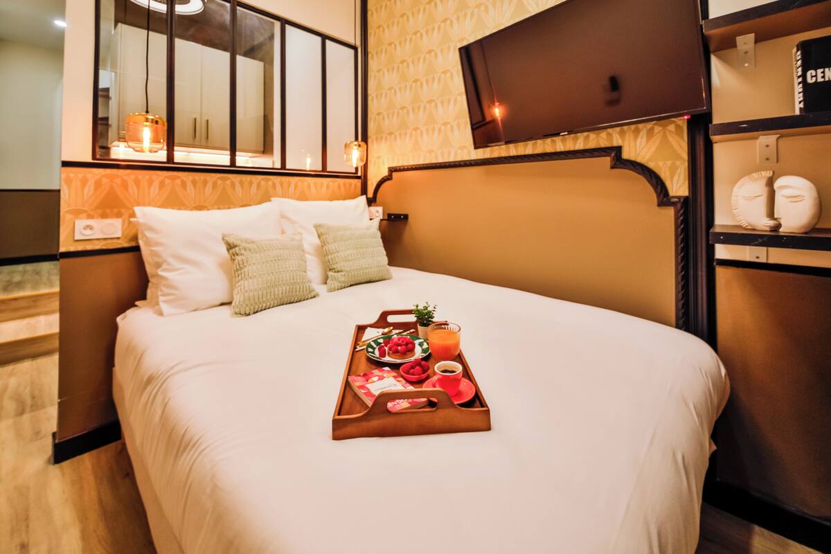 A neatly made bed with a tray of breakfast items, a television above the bed, and a patterned wall.