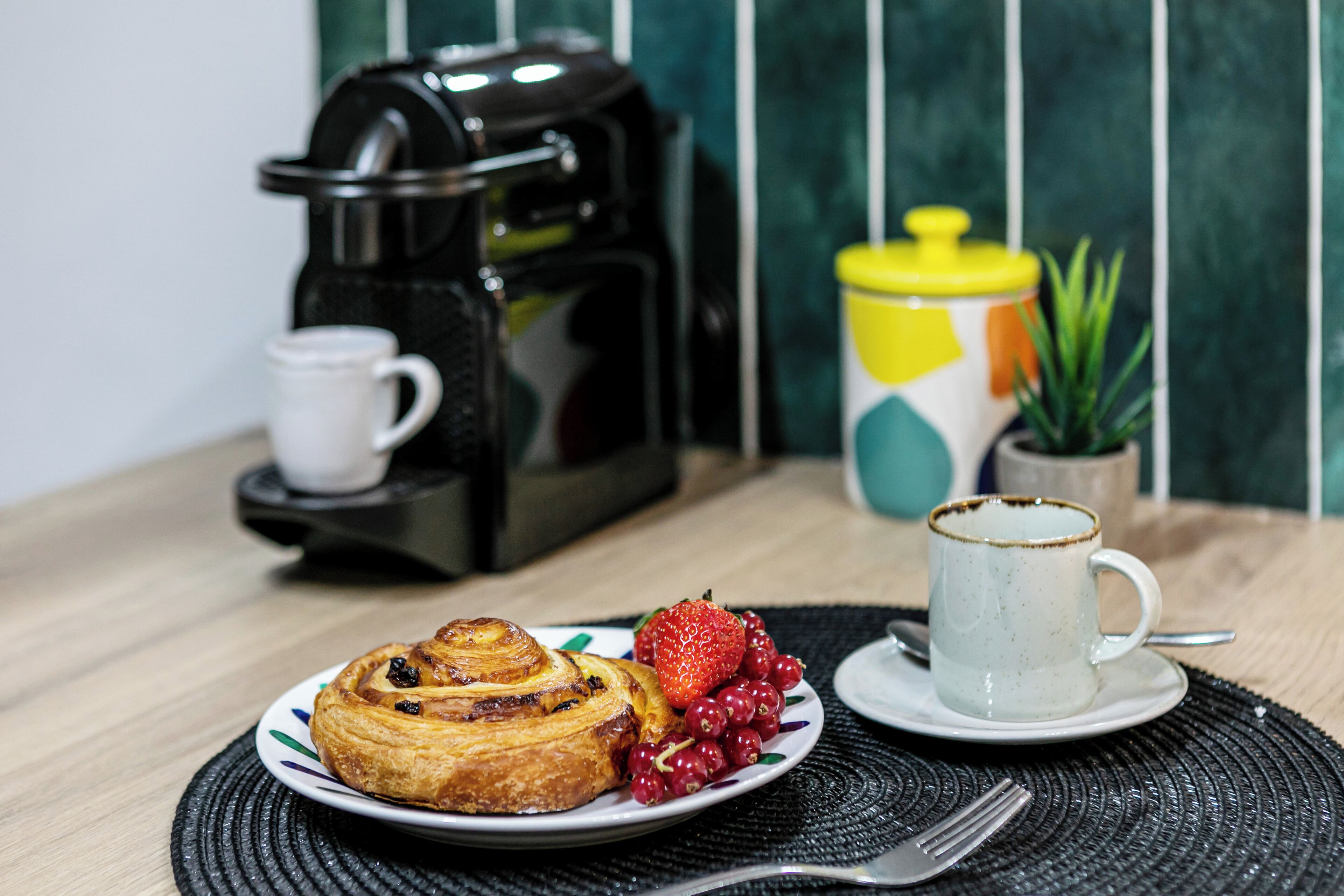 A breakfast setting with a pastry, fruit, a coffee maker, and a mug on a table.