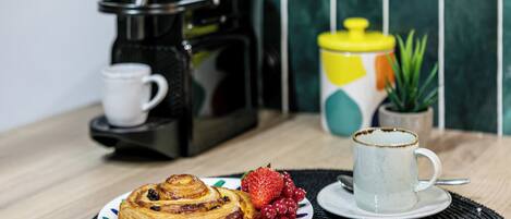 A breakfast setting with a pastry, fruit, a coffee maker, and a mug on a table.