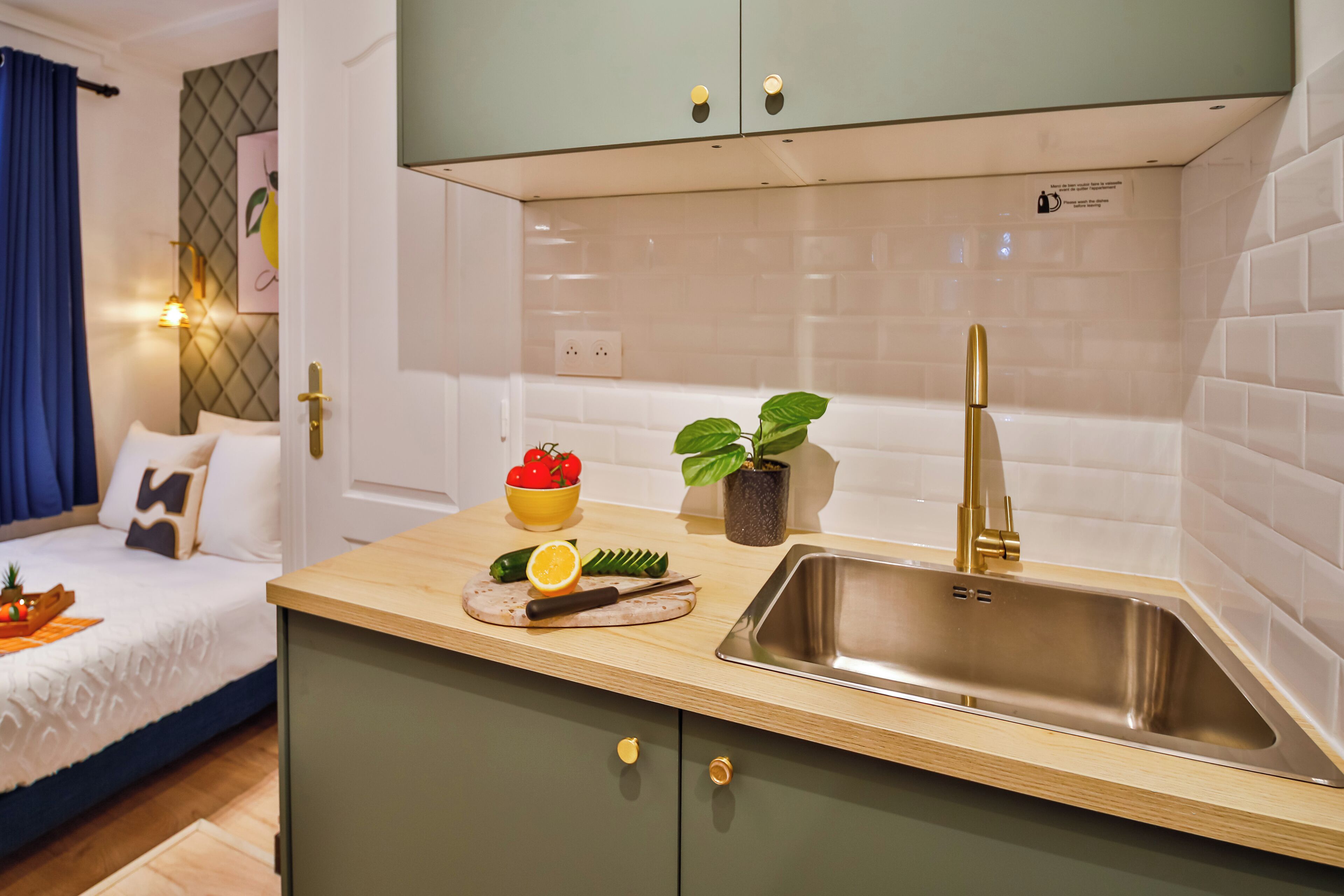 A compact kitchen with a sink, countertop, and green cabinets. There is a bed with a blue headboard and a potted plant on the counter.