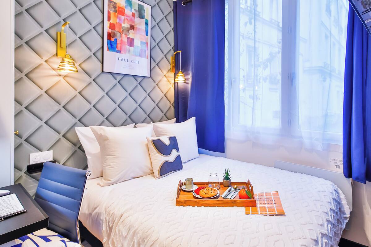 A neatly made bed with a tray of breakfast items, a desk with a laptop, and a wall with a geometric pattern and two wall-mounted lamps.