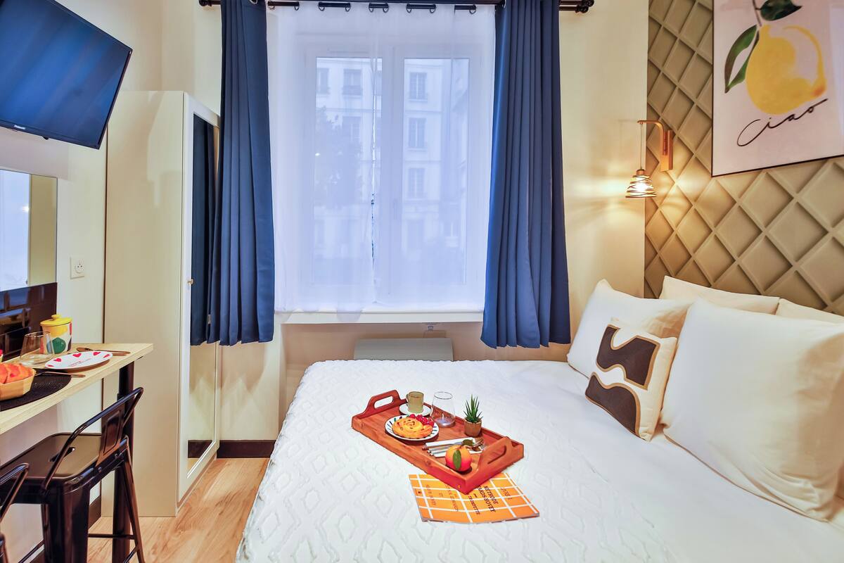 A hotel room with a bed, a desk with a chair, a tray with breakfast items, and a window with curtains.