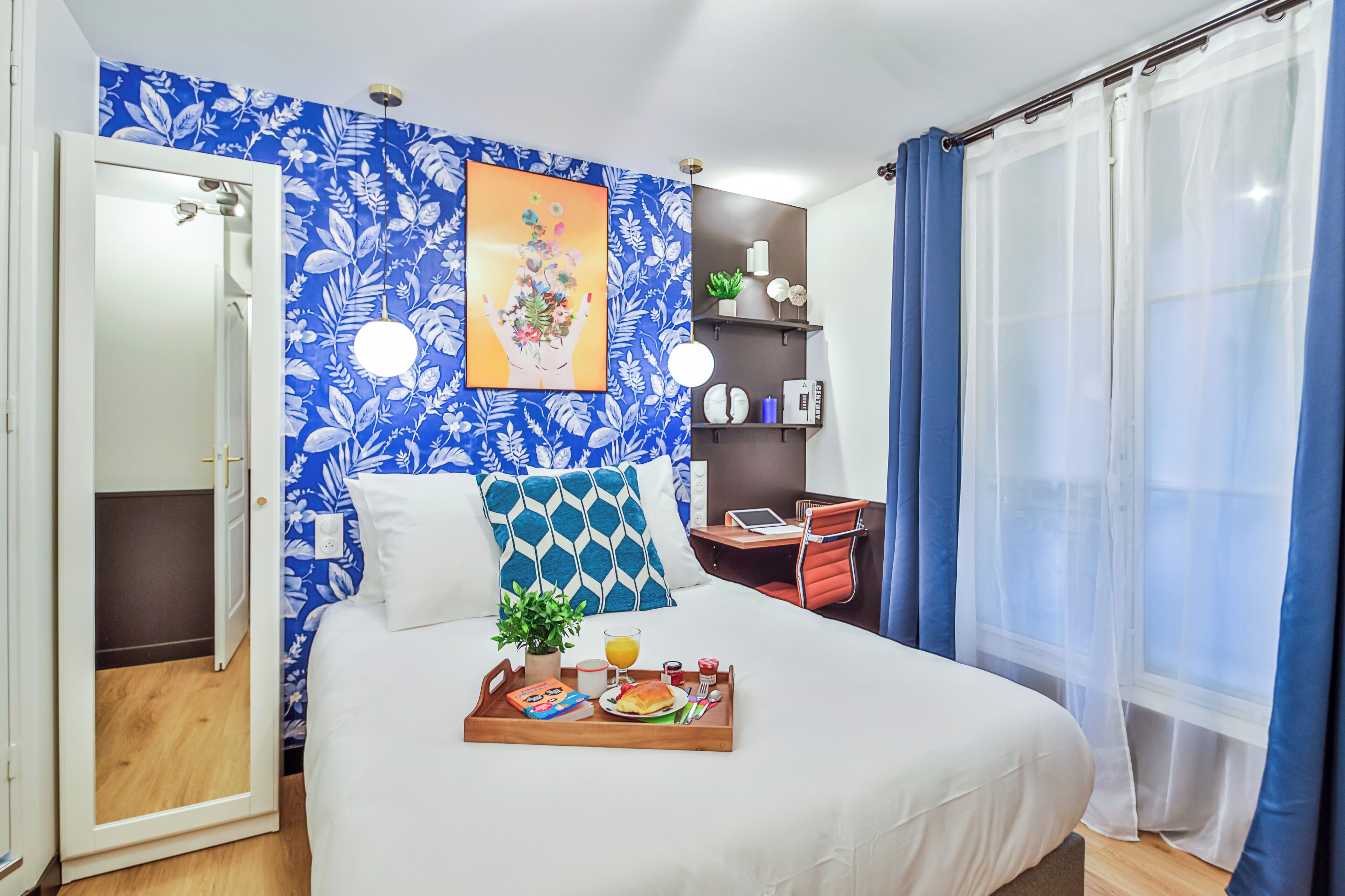 A hotel room with a bed, breakfast tray, a desk with a laptop, a small plant, and a window with curtains.